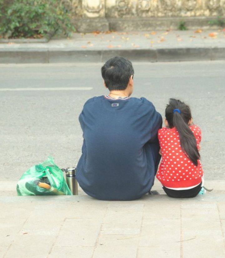 A father and his young daughter sitting together on a kerb, seen from behind